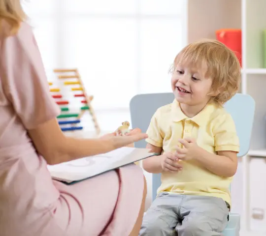 A woman in a pink dress sits across from a child in a yellow shirt, engaging them with a small toy in a colorful educational setting.