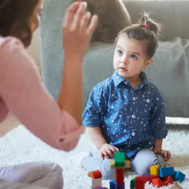 A child sits on a carpet, engaged in building with colorful blocks while an adult gestures animatedly in conversation.